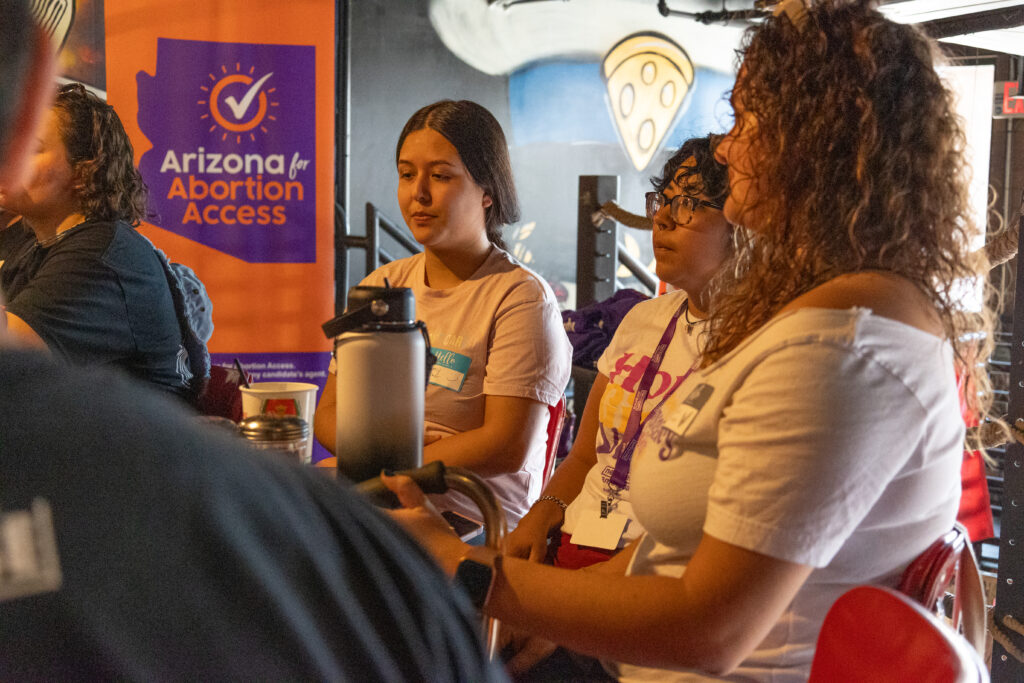 A group of three yes on prop 139 canvassers sitting at a table in front of an orange and purple Arizona for Abortion Access banner at Spinelli's Pizza in Tempe, AZ