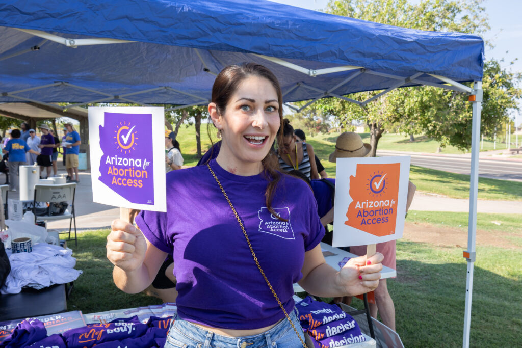 An enthusiastic yes on prop 139 volunteer holding up small Arizona for Abortion Access signs used as fans at Kiwanis Park in Tempe, AZ