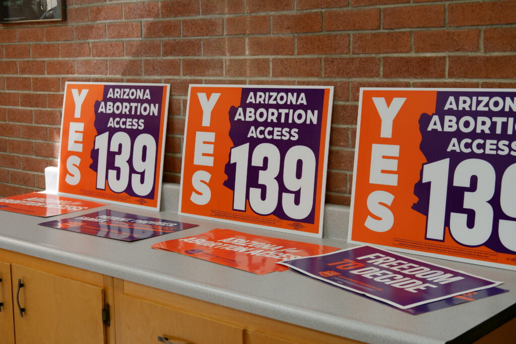 A row of orange and purple yes on prop 139 Arizona Abortion Access yard signs on a countertop near a brick wall