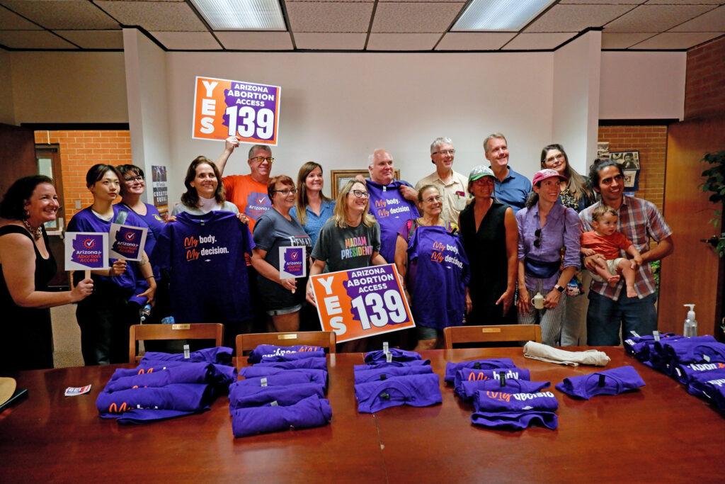 Large group of yes on prop 139 Arizona Abortion Access canvassers in Tucson holding signs in front of a wooden table filled with my body, my decision t-shirts