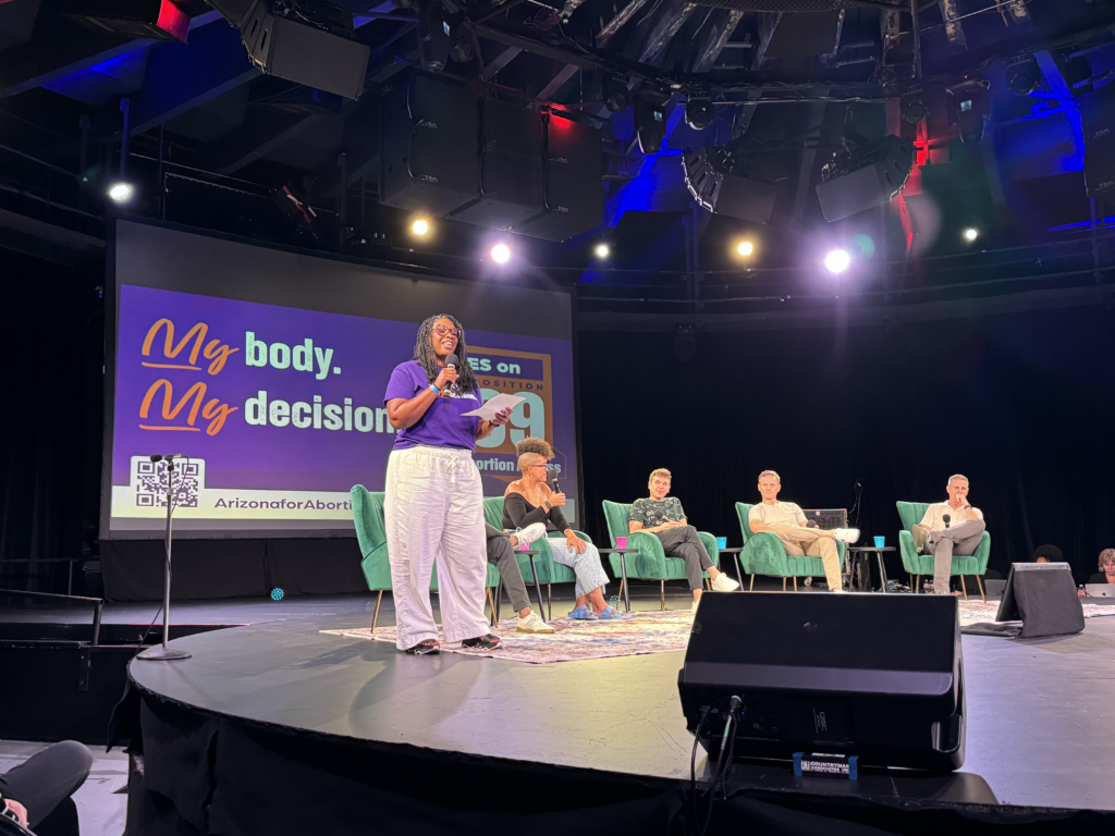 Chris Love on stage with Pod Save America in front of a large yes on prop 139 My Body, My Decision sign in front of a crowd in Phoenix, AZ