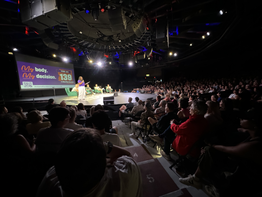 Chris Love on stage with Pod Save America in front of a large yes on prop 139 My Body, My Decision sign in front of a crowd in Phoenix, AZ