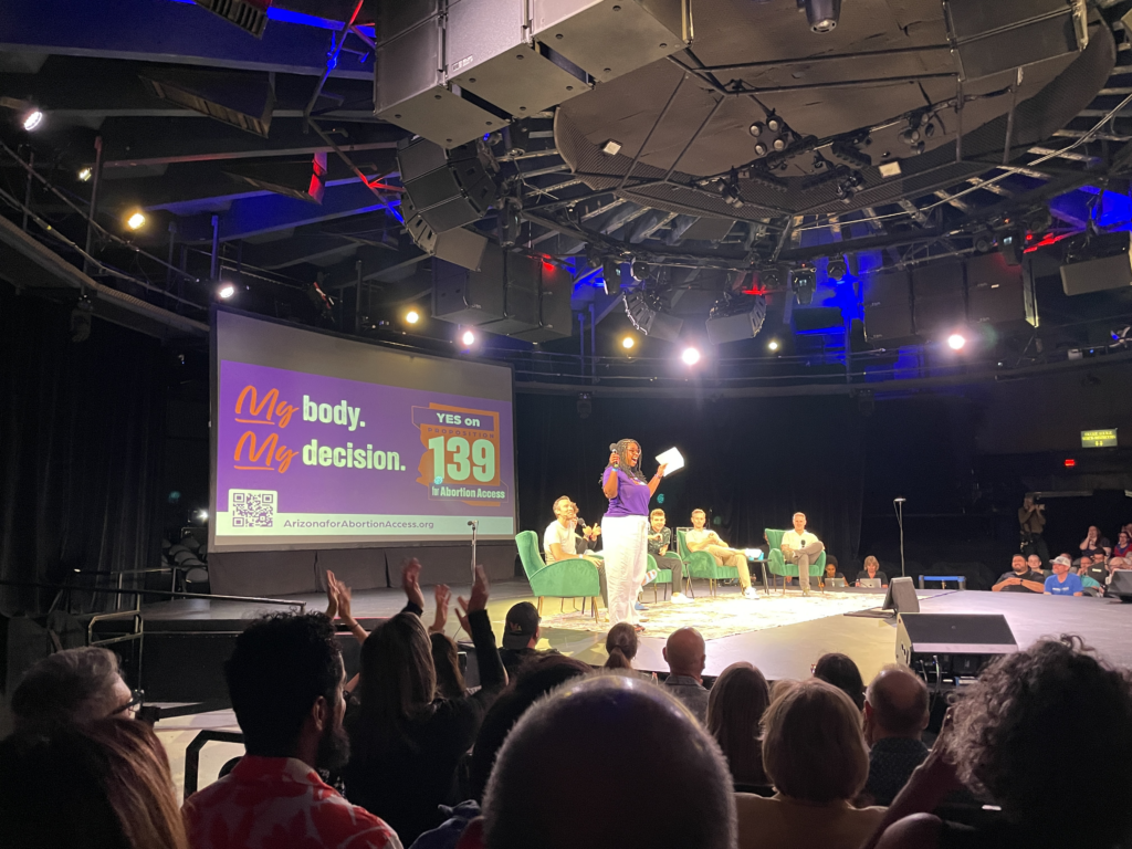 Chris Love on stage with Pod Save America in front of a large yes on prop 139 My Body, My Decision sign in front of a crowd in Phoenix, AZ