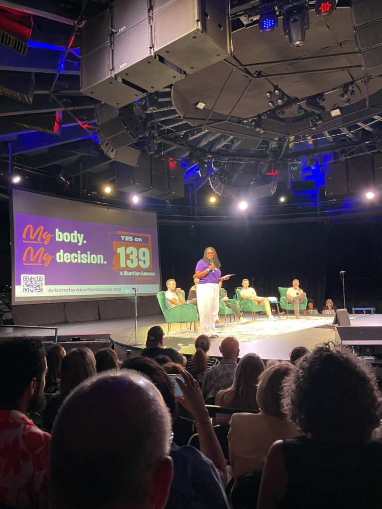 Chris Love on stage with Pod Save America in front of a large yes on prop 139 My Body, My Decision sign in front of a crowd in Phoenix, AZ